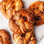Four golden-brown soft pretzels topped with coarse salt are arranged on parchment paper, with a small dish of salt nearby.