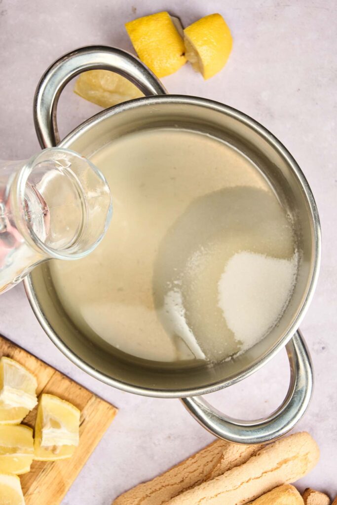 Lemon juice is poured into a saucepan with sugar to make a simple syrup, with sliced lemons and ladyfingers nearby.