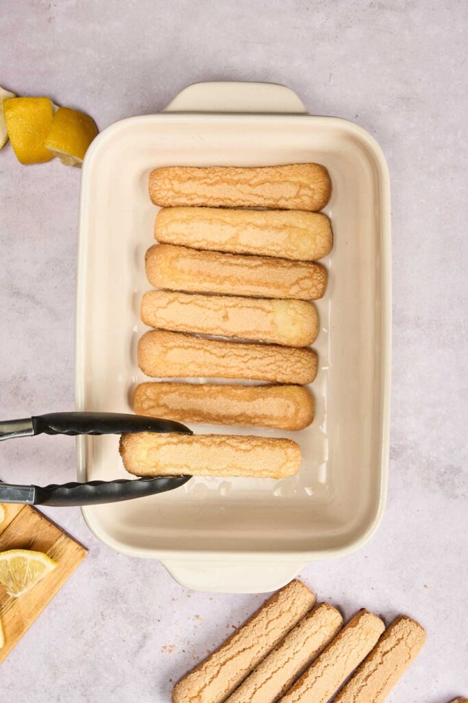 Tongs placing a ladyfinger cookie into a white rectangular baking dish with several ladyfingers already arranged inside, perfect for assembling a refreshing Lemon Tiramisu.