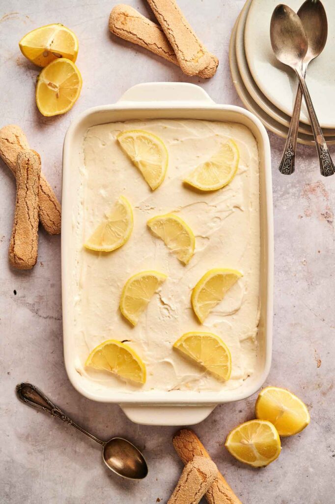 Overhead shot of lemon tiramisu topped with fresh lemon wedges in a white baking dish.