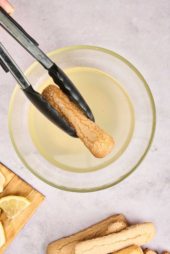 A pair of tongs holds a ladyfinger over a bowl of liquid, with more ladyfingers and lemon slices on a cutting board&mdash;ingredients ready for crafting a refreshing Lemon Tiramisu.