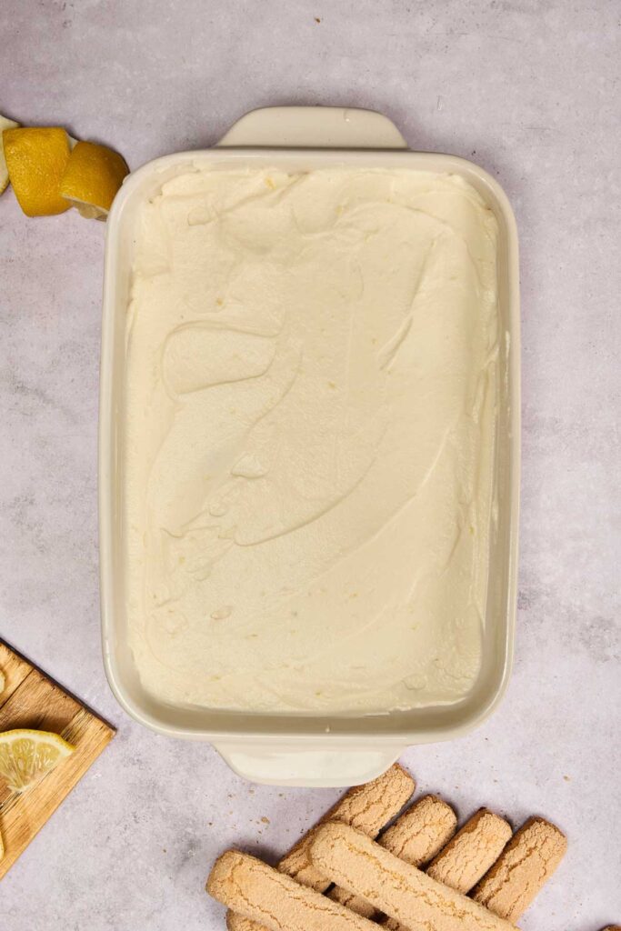 A rectangular baking dish filled with a smooth, creamy Lemon Tiramisu layer, surrounded by lemon slices and ladyfinger cookies on a light surface.