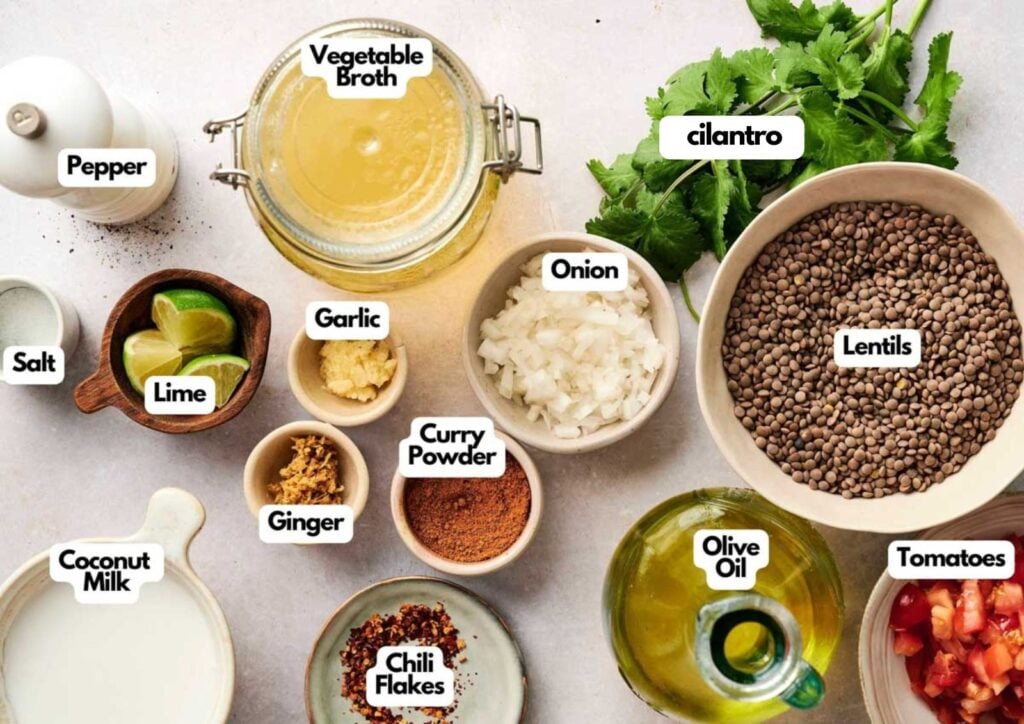 Top view of ingredients in bowls and jars for Lentil Soup dish, including lentils, onion, tomatoes, garlic, spices, olive oil, coconut milk, lime, and cilantro—all labeled.