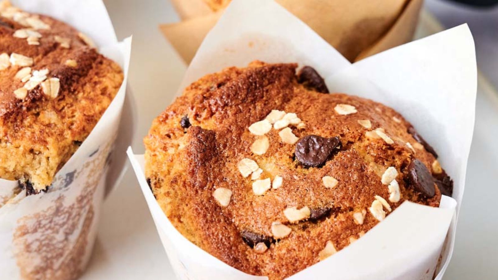 A close-up of a muffin topped with oats and chocolate chips, wrapped in white parchment paper.