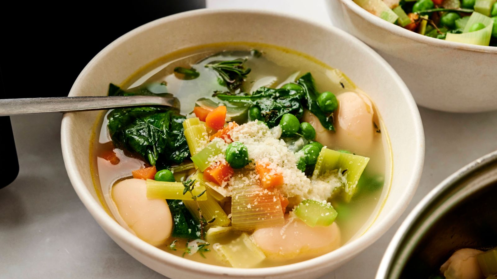 A bowl of vegetable soup with spinach, peas, carrots, celery, and large white beans, topped with grated cheese.