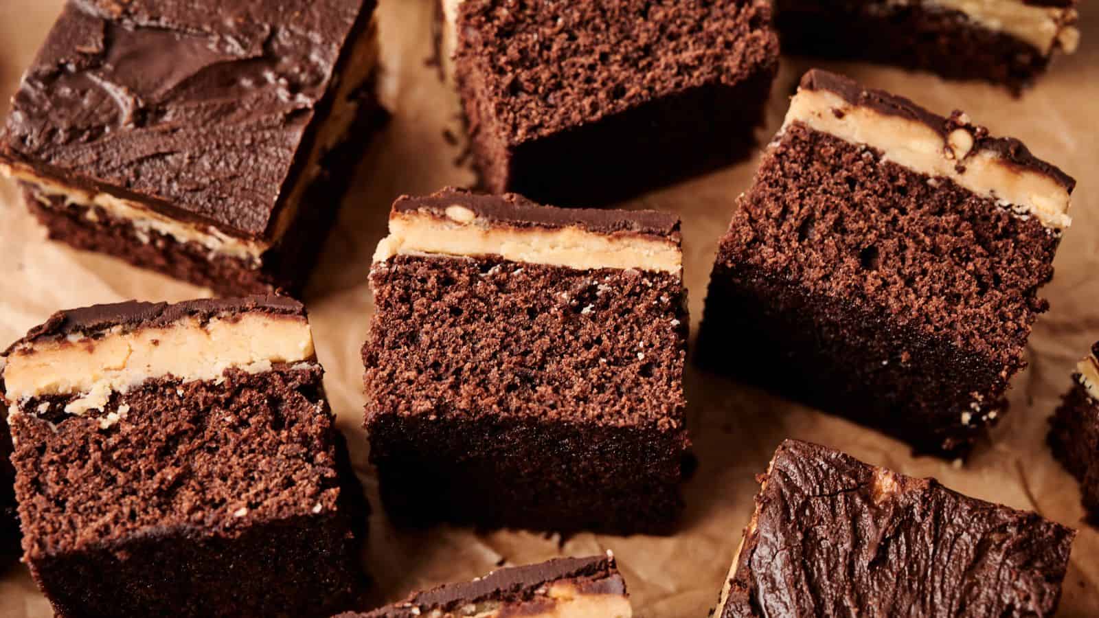 Several squares of chocolate cake with a layer of peanut butter filling and a chocolate topping are arranged on brown parchment paper.