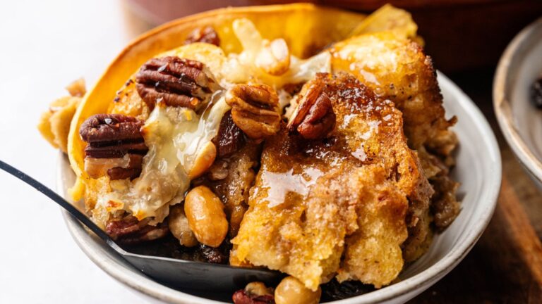 A bowl of bread pudding topped with pecans, peanuts, melted butter, and syrup, with a black spoon on the side.