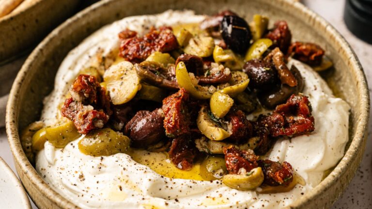 A bowl of whipped feta cheese topped with mixed olives, sun-dried tomatoes, olive oil, and cracked black pepper.