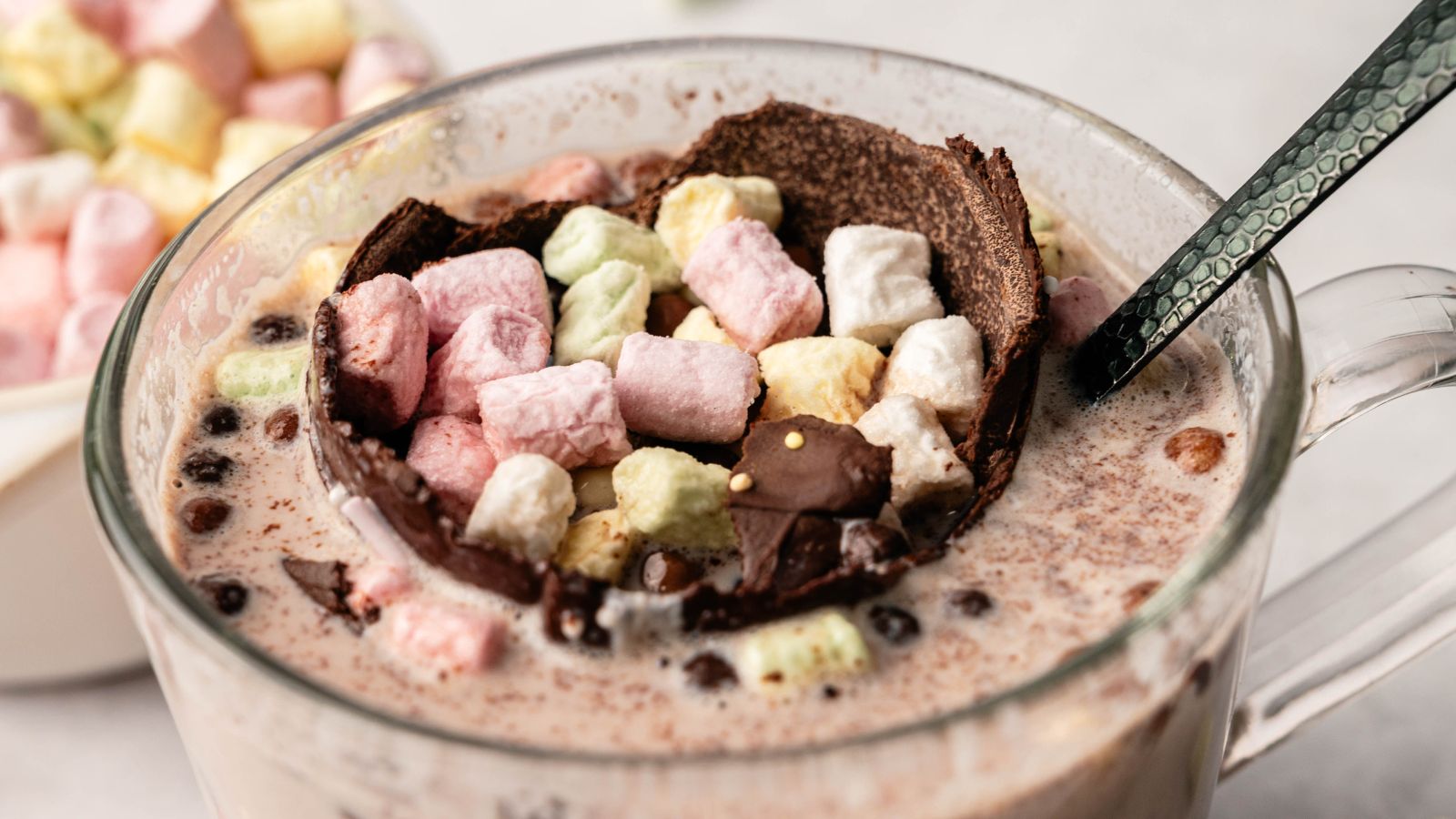 A glass mug filled with hot chocolate, topped with a chocolate shell and colorful mini marshmallows, with a spoon resting inside.