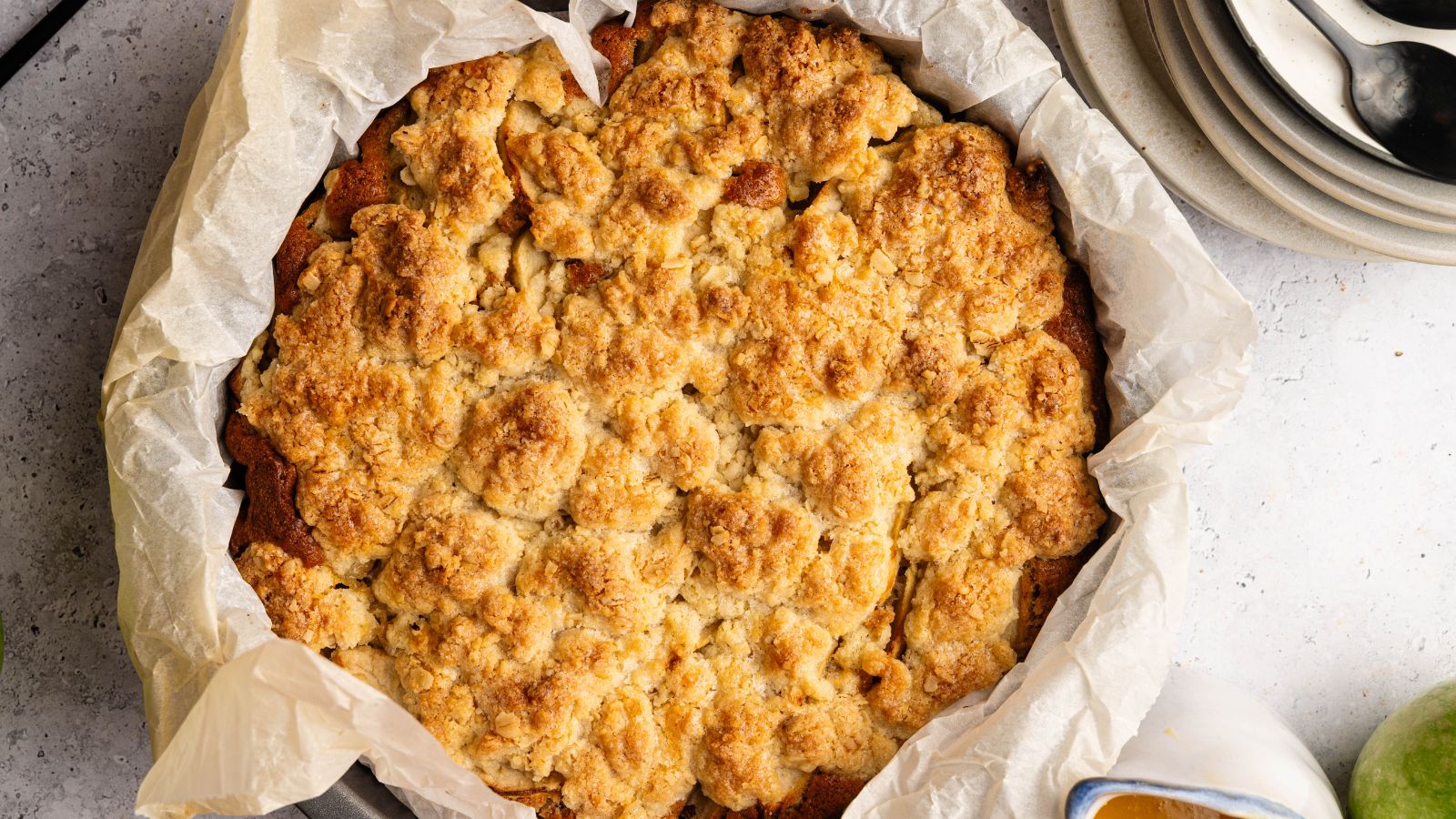 A baked apple crumble sits in a round, parchment-lined pan next to a stack of plates and spoons on a light countertop.