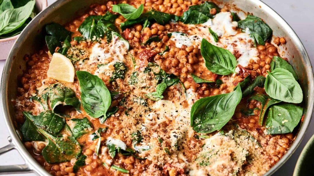 A pan of lentil dish with spinach, fresh basil, tomato sauce, and melted cheese, garnished with lemon wedges.