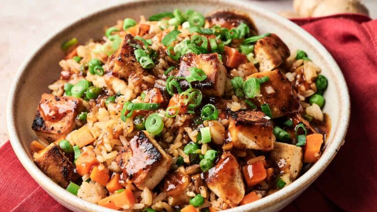 A bowl of fried rice topped with grilled tofu cubes, diced carrots, peas, and chopped green onions, garnished with a dark sauce.