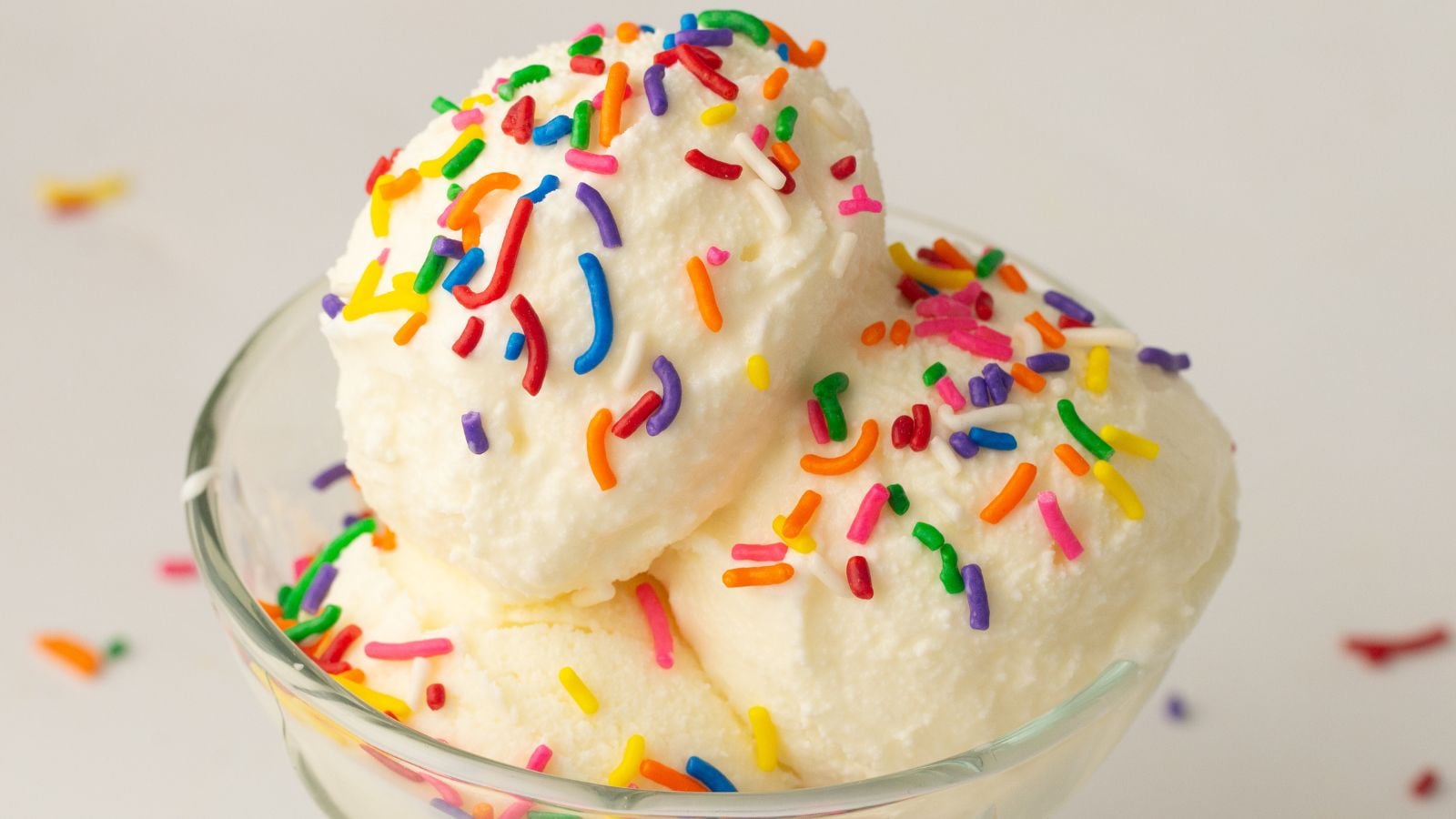 A glass bowl with two scoops of vanilla ice cream topped with colorful rainbow sprinkles, set against a plain white background.