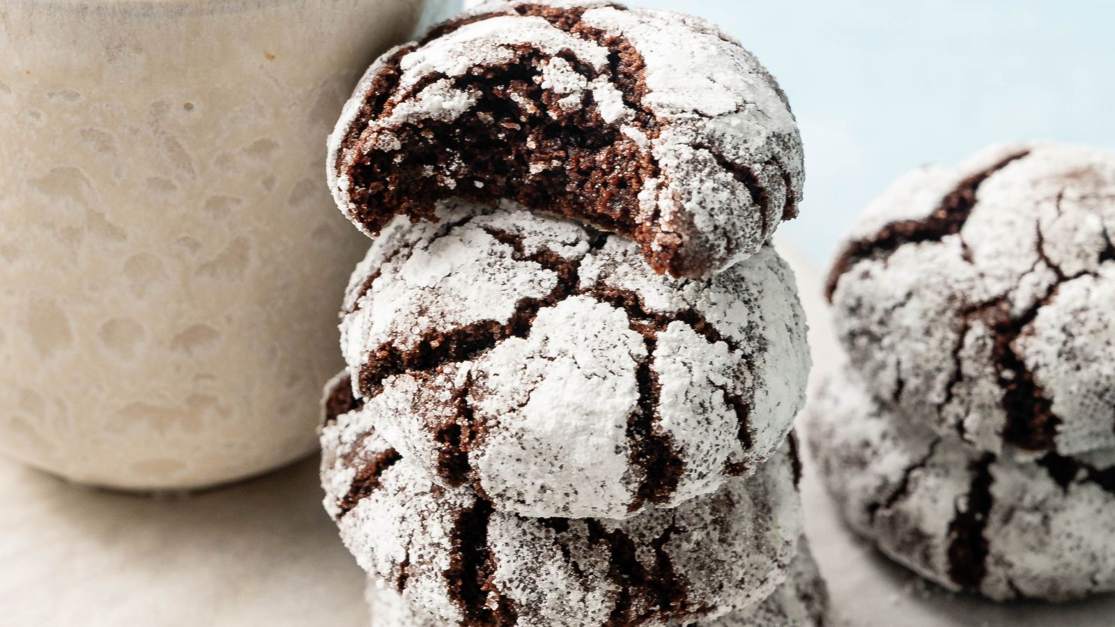 A stack of powdered sugar-coated chocolate crinkle cookies, with one cookie on top showing a bite taken out, next to a light-colored cup.