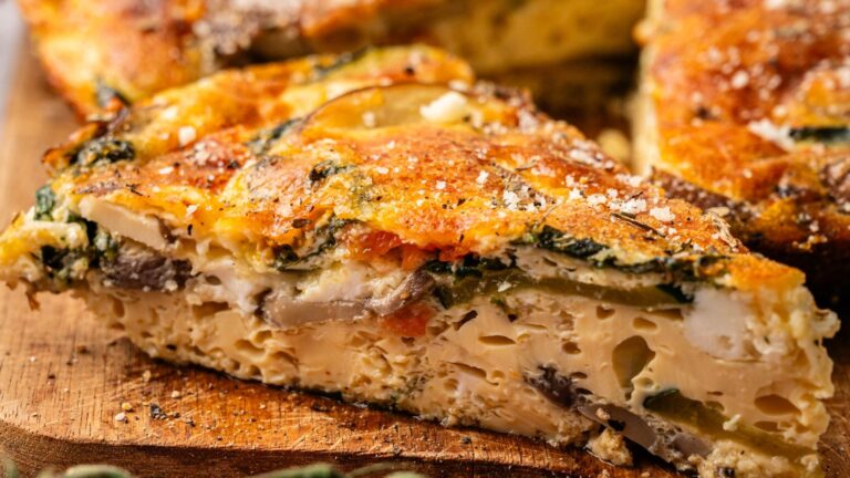 A close-up of a slice of vegetable frittata with visible layers of eggs, mushrooms, spinach, and melted cheese on a wooden surface.