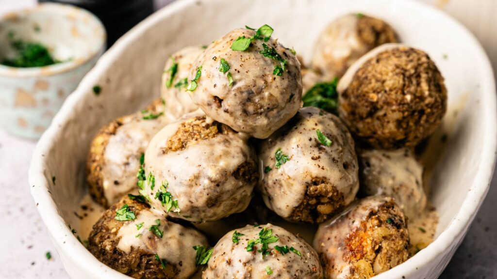 A white bowl filled with several meatballs topped with a creamy sauce and garnished with chopped parsley.