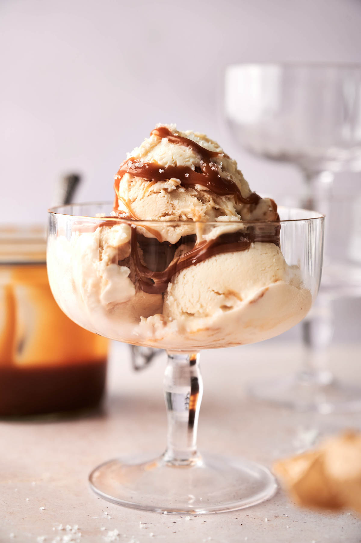 A glass dessert dish filled with two scoops of creamy No Churn Ice Cream topped with caramel sauce, with blurred glassware and a jar in the background.
