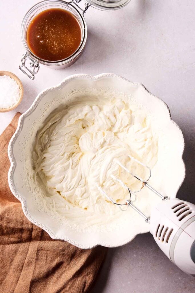 A hand mixer whips cream in a white bowl, the first step for making No-Churn Salted Caramel Ice Cream. Nearby are a jar of caramel sauce, a small bowl of flaky salt, and a brown cloth on a light countertop.