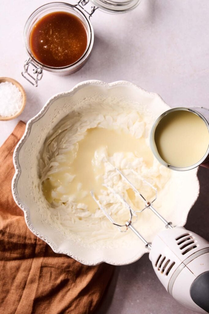 A hand-held mixer whips cream in a white bowl as sweetened condensed milk is poured in—the perfect base for No-Churn Salted Caramel Ice Cream; two jars and a small bowl are nearby.
