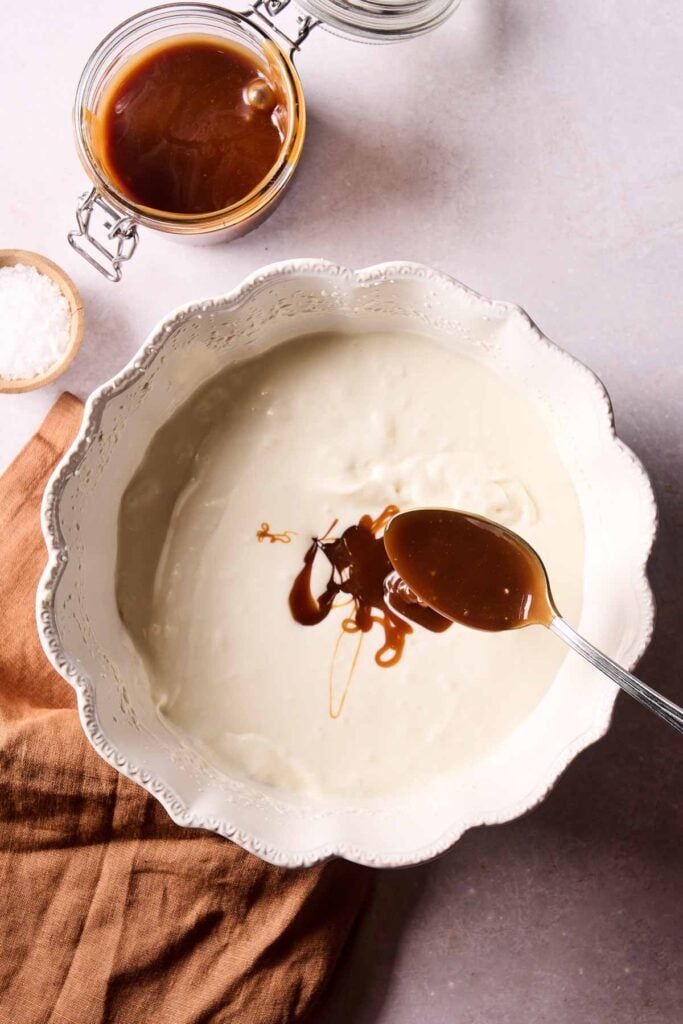 A spoon drizzles caramel sauce over a bowl of white batter, the first step in making creamy No-Churn Salted Caramel Ice Cream. An open jar of caramel and a small bowl of salt are nearby on a light surface, ready to add that irresistible sweet and salty finish.