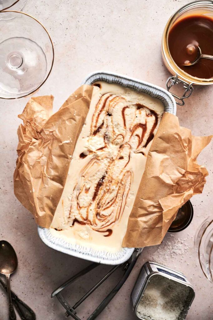 A rectangular pan lined with brown parchment paper holds swirled No Churn Ice Cream; caramel sauce, spoons, and glassware are arranged nearby on a light surface.