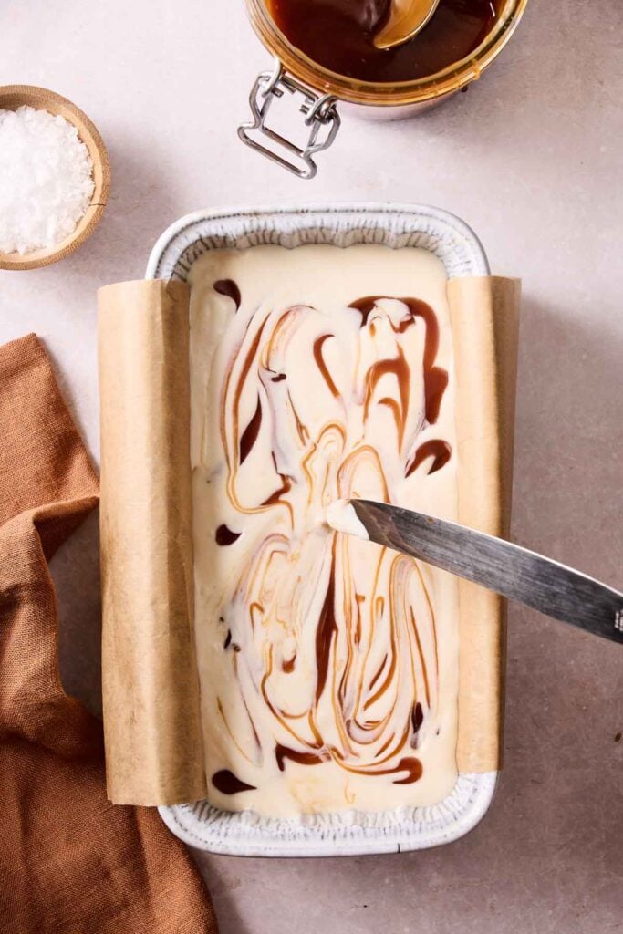 A loaf pan lined with parchment paper holds swirled No-Churn Salted Caramel Ice Cream and caramel sauce. A knife rests on top. A bowl of caramel sauce and a dish of salt are nearby.