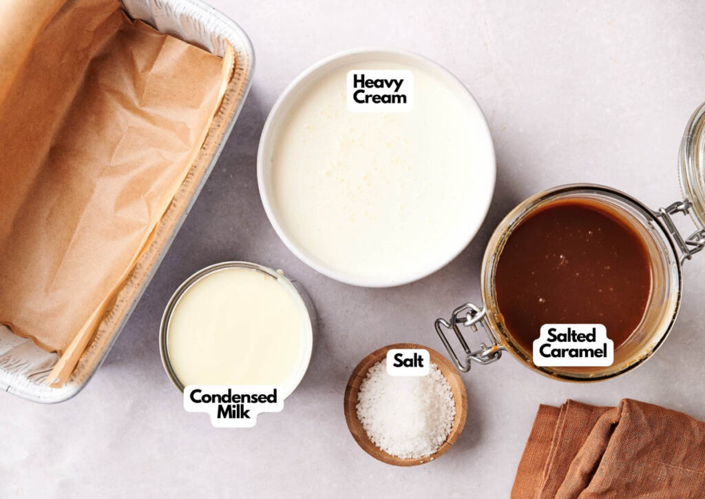 A parchment-lined pan, a bowl of heavy cream, a jar of salted caramel, an open can of condensed milk, and a small bowl of salt are arranged on a countertop&mdash;everything you need to make creamy No Churn Ice Cream.