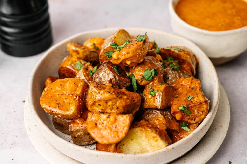 A bowl of Patatas Bravas&mdash;crispy roasted potato wedges topped with a creamy orange sauce and garnished with chopped herbs, with a bowl of extra sauce in the background.