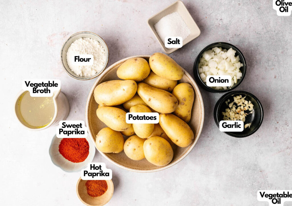 A bowl of whole potatoes for Patatas Bravas is surrounded by small bowls of flour, vegetable broth, sweet paprika, hot paprika, salt, chopped onion, chopped garlic, vegetable oil, and olive oil.