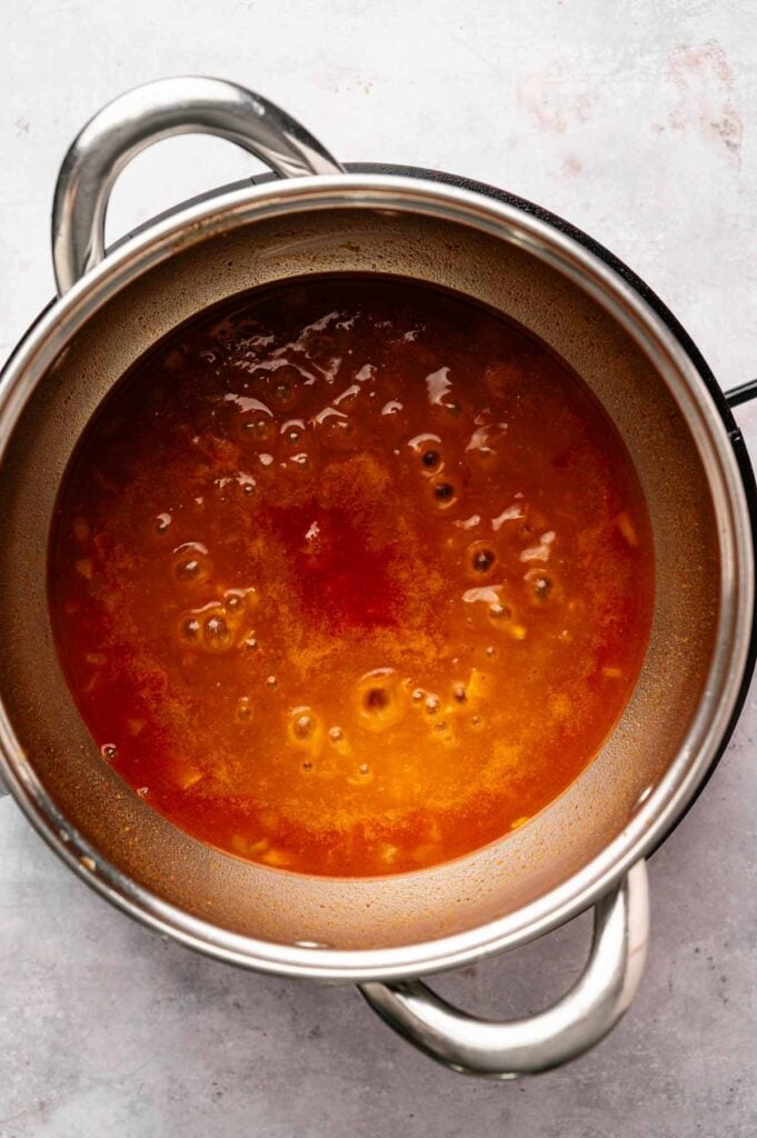 A stainless steel pot filled with simmering orange-red broth, reminiscent of the bold flavors in Patatas Bravas, sits on a light-colored surface.