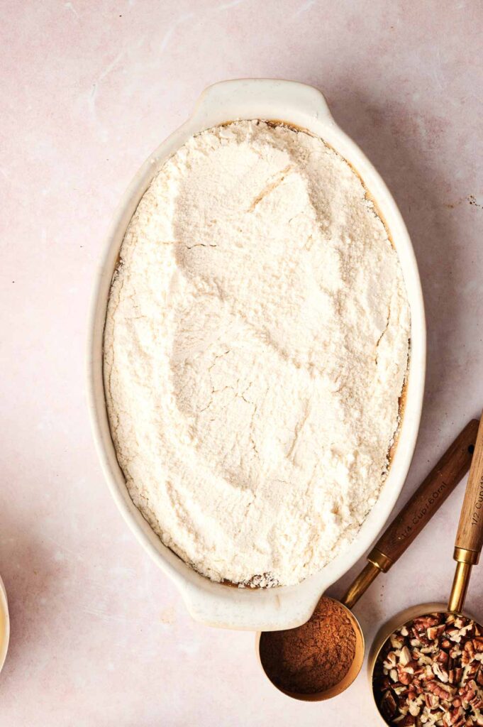Oval baking dish filled with an even layer of flour mixture, surrounded by a measuring cup of cinnamon, chopped pecans, and two wooden-handled utensils on a light countertop—perfect for preparing an Air Fryer Pumpkin Dump Cake.