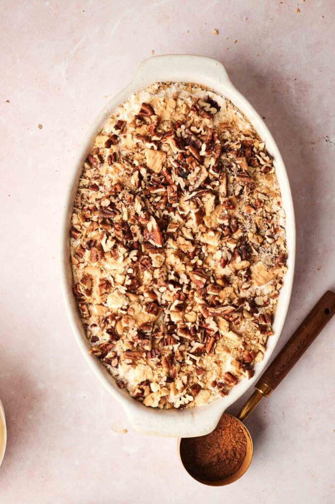 Oval baking dish filled with a crumbly pecan topping, resting on a light surface with a measuring spoon beside it—perfect for serving Air Fryer Pumpkin Dump Cake.
