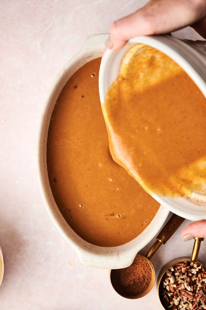 A person pours a thick, pumpkin mixture into a large oval baking dish on a light surface with spices and chopped nuts nearby, preparing the base for an Air Fryer Pumpkin Dump Cake.