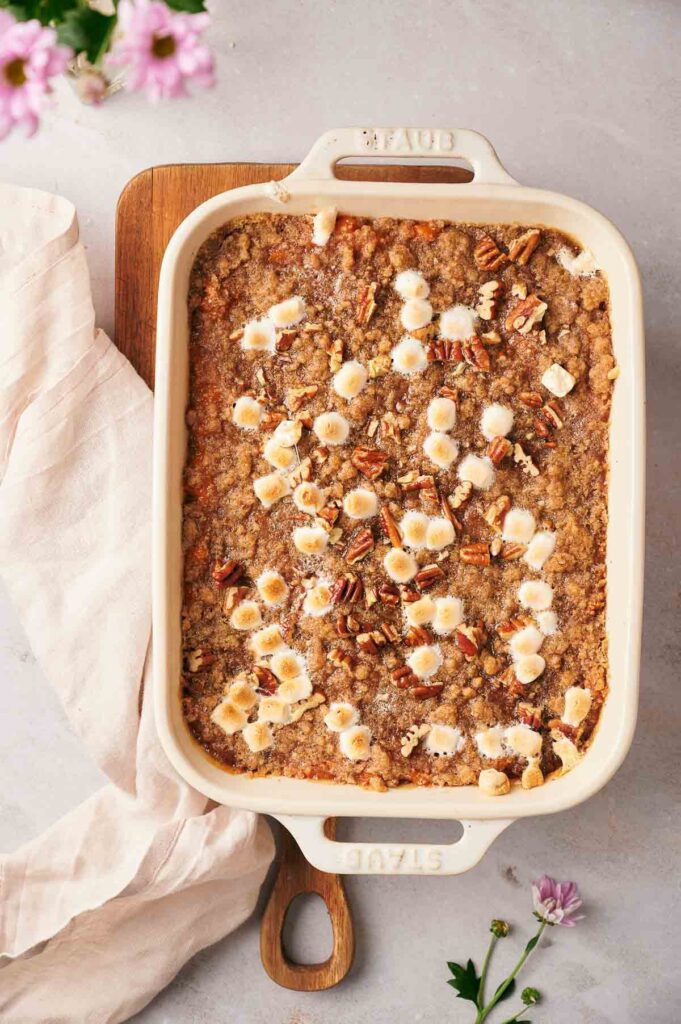 A Sweet Potato Casserole topped with toasted marshmallows and chopped pecans, baked in a white rectangular dish and served on a wooden board.
