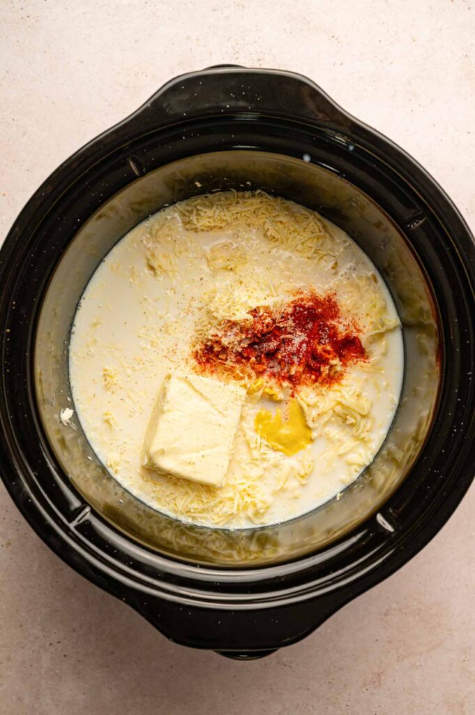 A slow cooker filled with uncooked macaroni, milk, shredded cheese, a block of butter, mustard, and paprika sets the stage for a delicious Slow Cooker Mac and Cheese before everything is mixed together.