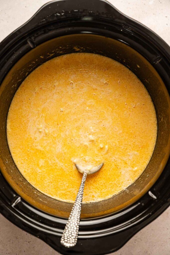 A spoon stirs a slow cooker filled with uncooked pasta, cheese, and milk, the mixture looking loose before it thickens.