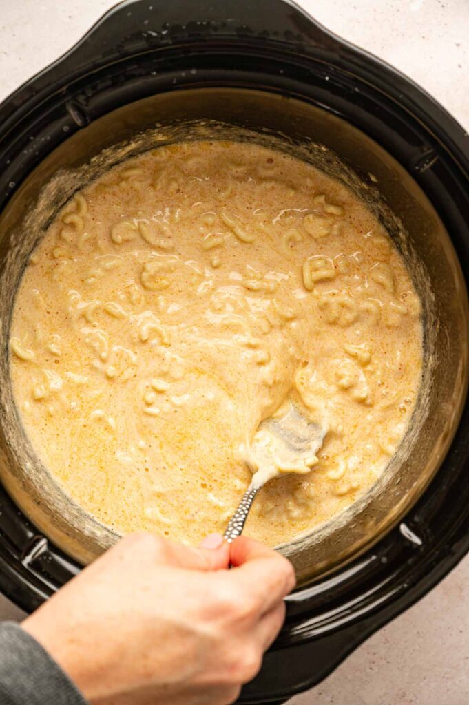 A hand stirs creamy Slow Cooker Mac and Cheese mixture in a slow cooker with a metal spoon.