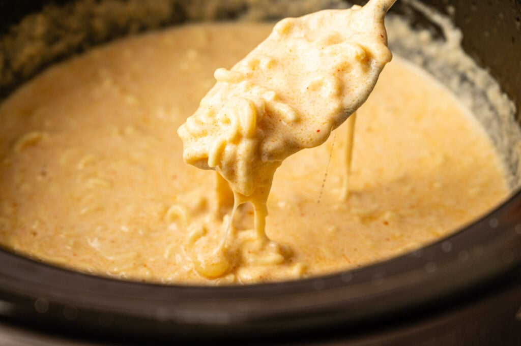 A spoon lifts creamy Slow Cooker Mac and Cheese from a pot, showing melted cheese and pasta in a thick, luscious sauce.