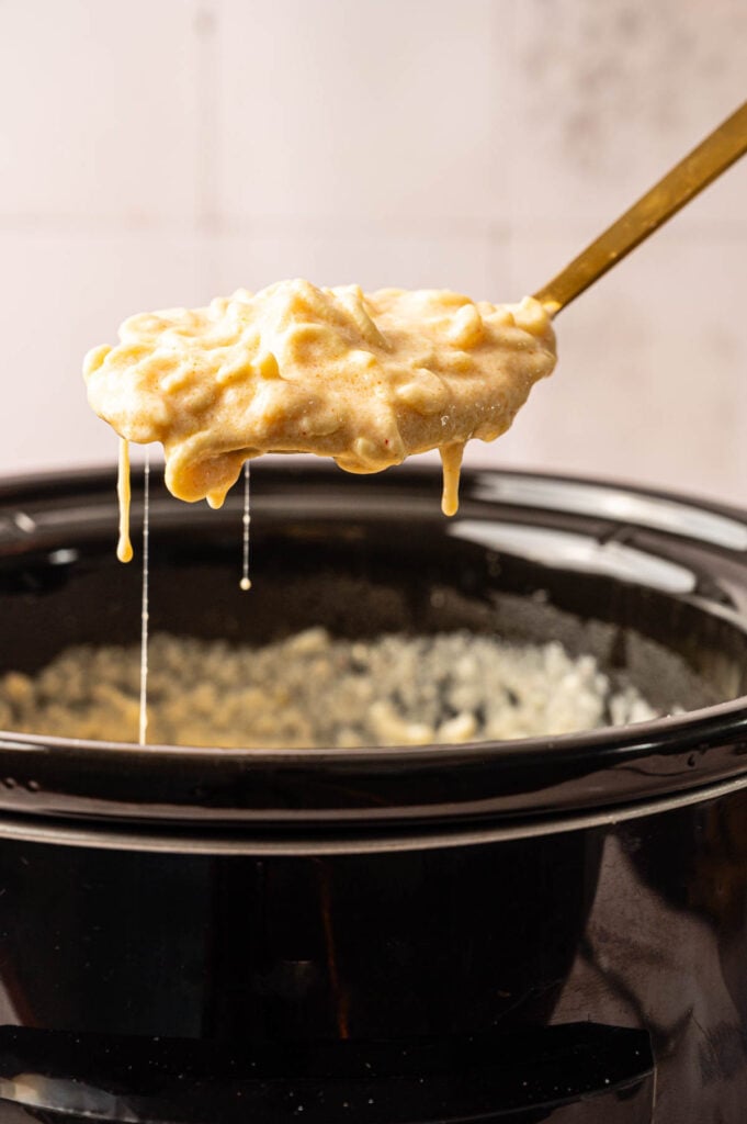 A spoon holds up creamy Slow Cooker Mac and Cheese above a black slow cooker, with cheese sauce dripping back into the pot.
