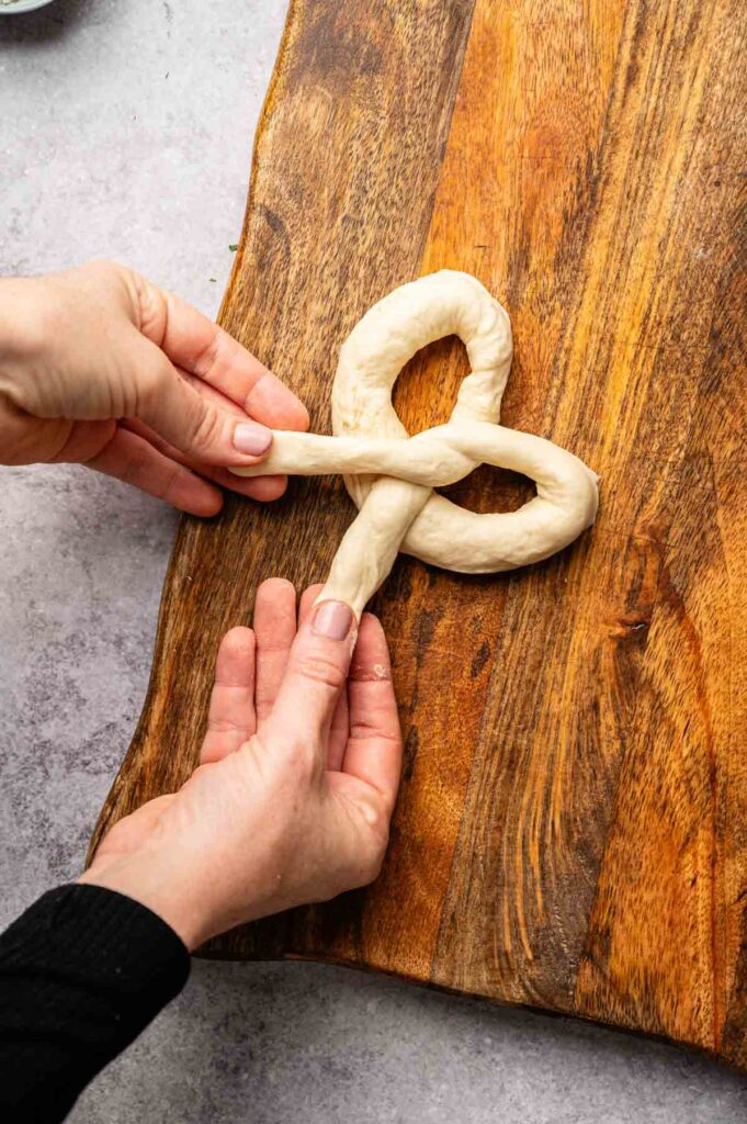 Two hands shape a piece of dough into a classic soft pretzel form on a wooden board.