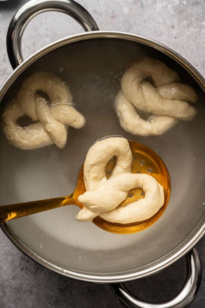 Three unbaked soft pretzels are being boiled in a pot of water, with one soft pretzel being lifted by a slotted spoon.