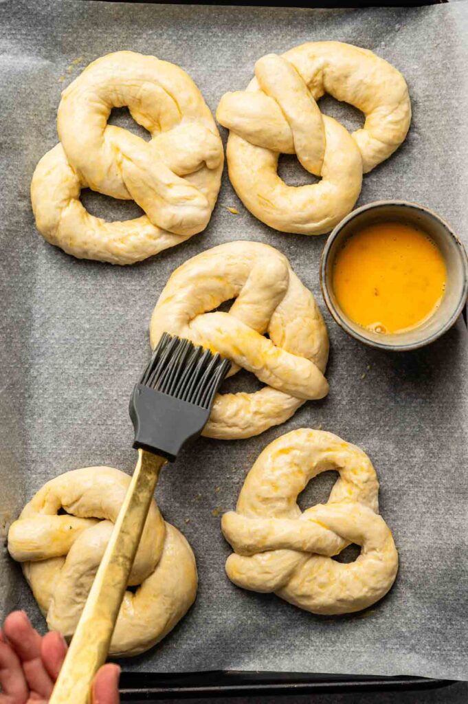 Six soft pretzels sit unbaked on a parchment-lined baking sheet, with a hand brushing egg wash on one; a small bowl of egg wash is nearby.