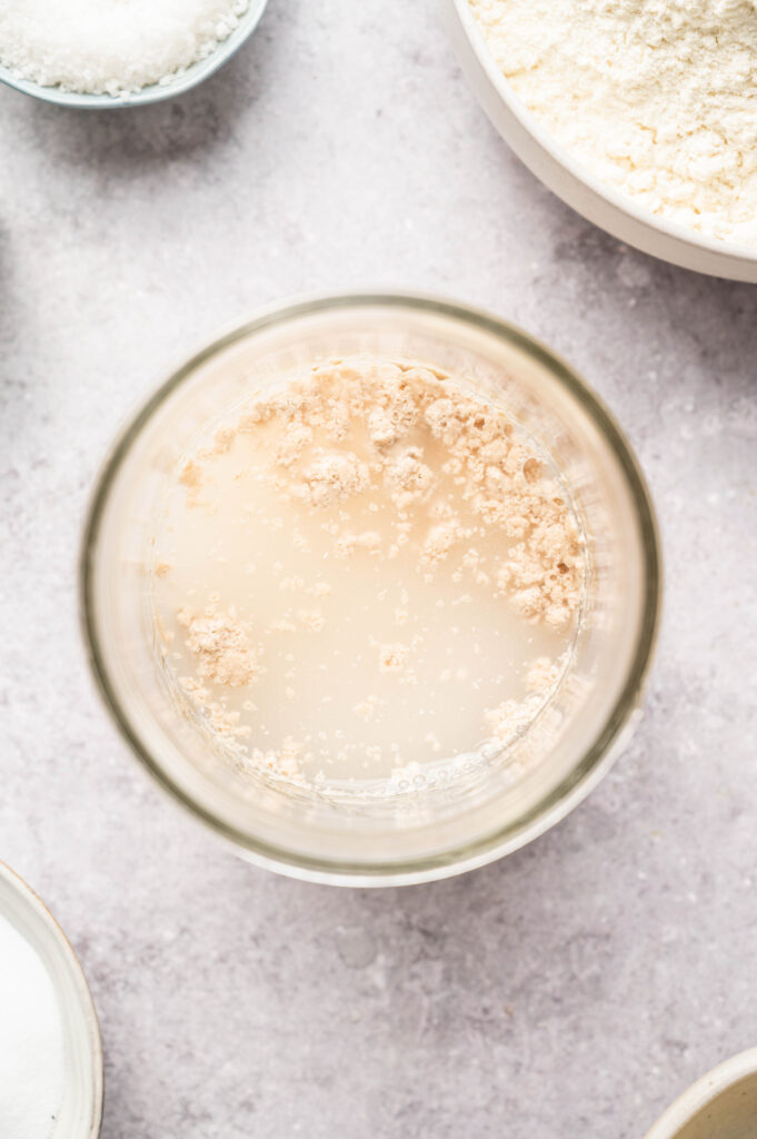 Yeast, sugar, and warm water sit in a glass bowl, starting to activate before turning foamy.