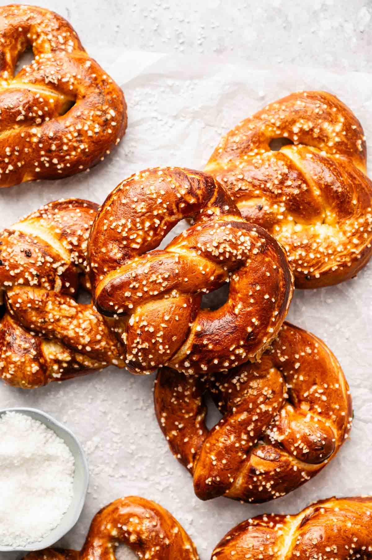 Several golden brown soft pretzels topped with coarse salt are arranged on a sheet of parchment paper, with a small bowl of salt nearby&mdash;perfect for anyone craving classic Soft Pretzels.