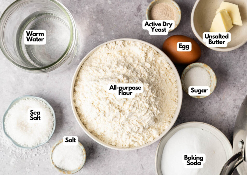 Top-down view of labeled baking ingredients in bowls on a countertop, including all you need to make delicious Soft Pretzels—flour, yeast, butter, egg, sugar, salt, baking soda, sea salt—and a glass of warm water.
