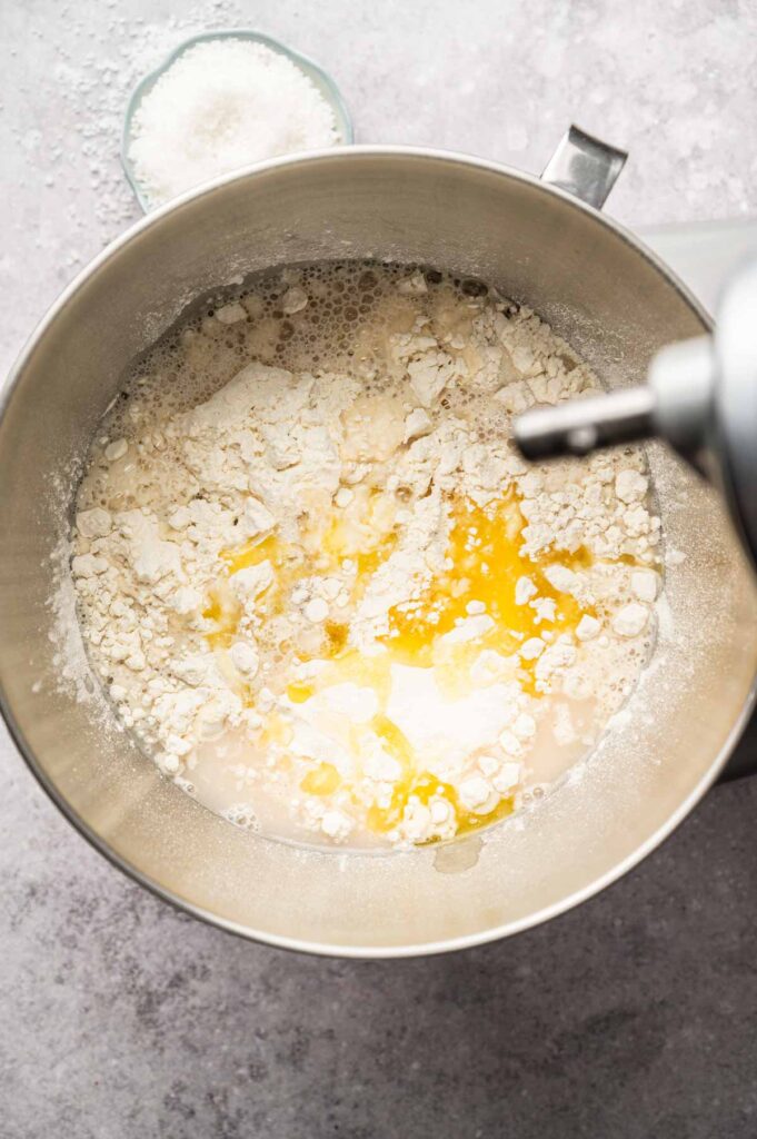 A stand mixer bowl contains flour, water, oil, and other ingredients needed for making soft pretzels before mixing. A small dish of salt is placed nearby on the countertop.