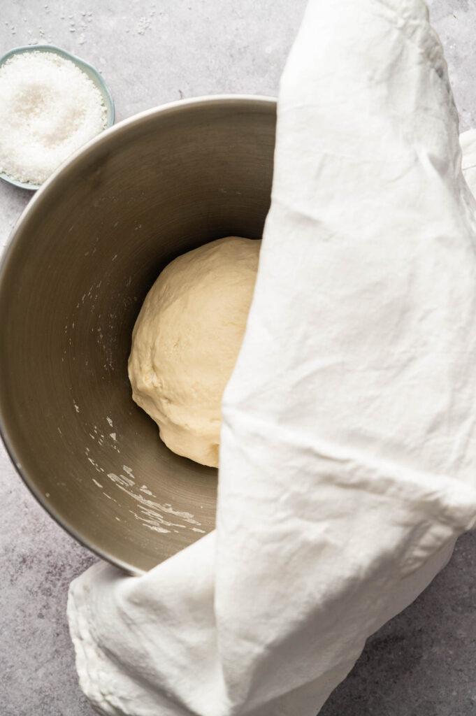 A metal bowl with dough for Soft Pretzels inside is partially covered by a white cloth. A small bowl of coarse salt is in the background.