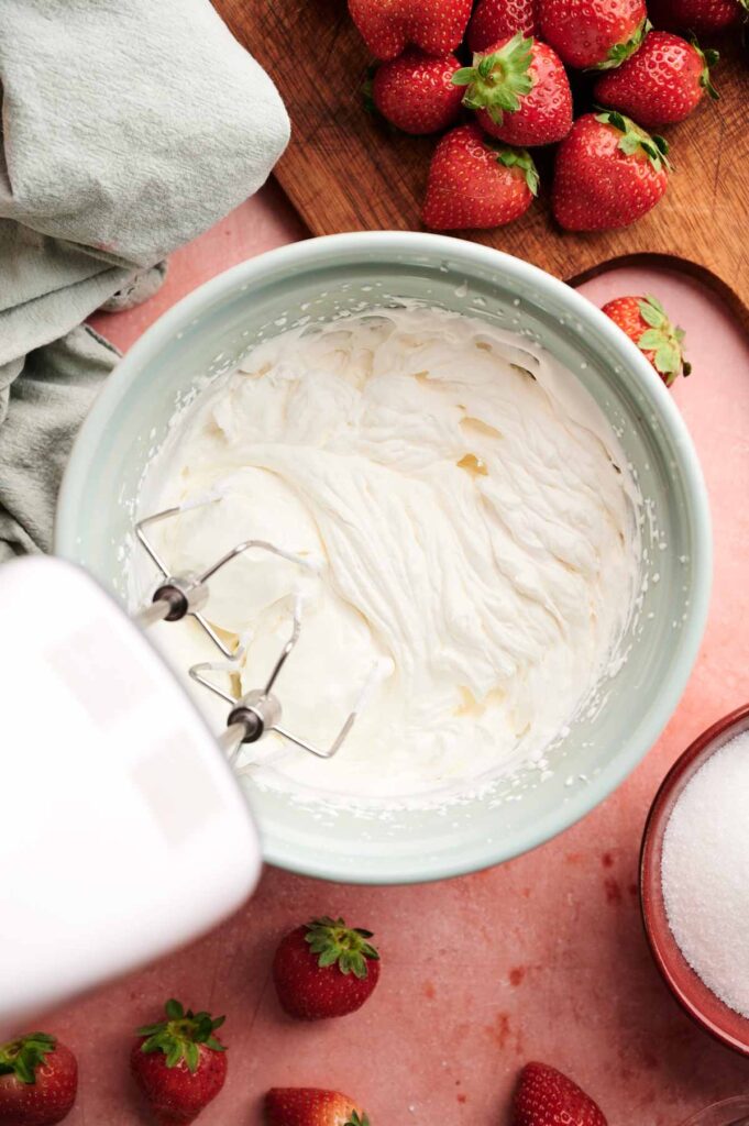 A hand mixer whips cream in a bowl on a pink surface, surrounded by fresh strawberries, a towel, and a bowl of sugar.