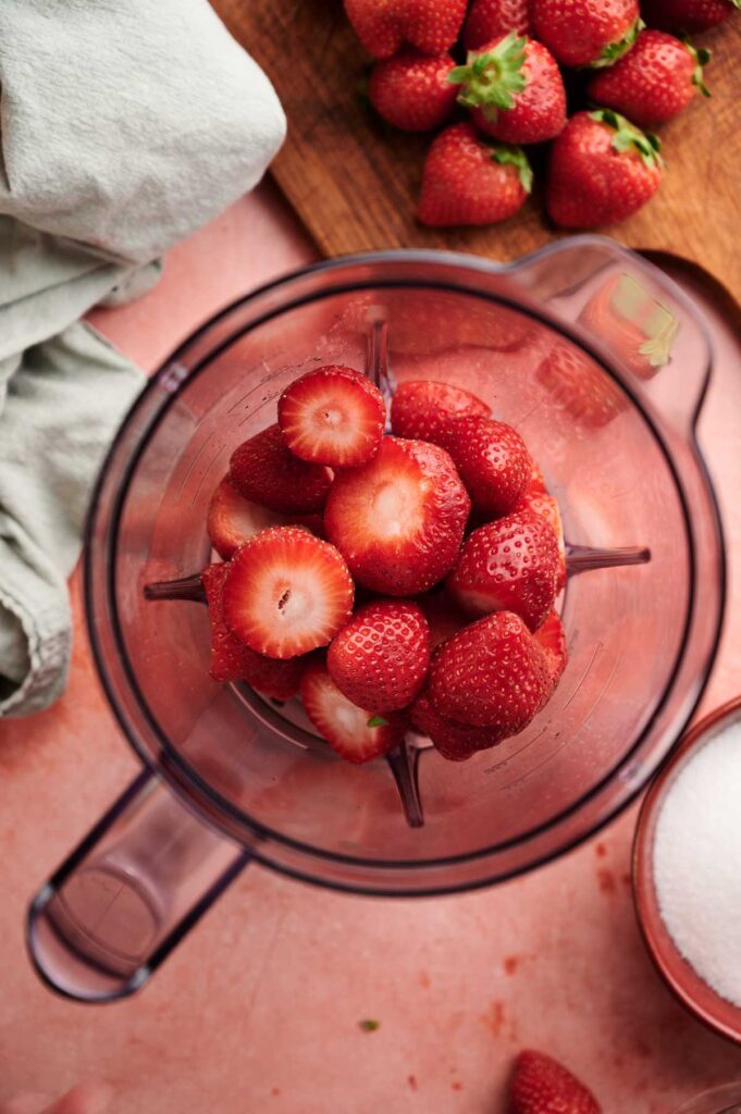 Overhead shot of hulled strawberries in a blender jar, with sugar and whole berries nearby.