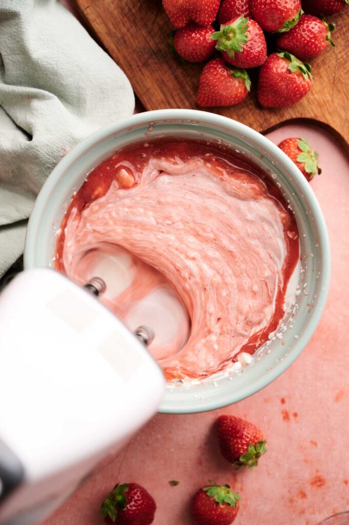 A hand mixer blends pink strawberry mixture in a bowl. Fresh strawberries are on a nearby cutting board and counter.